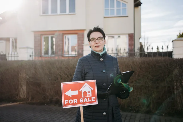 Woman realtor with eyeglasses holding a clipboard and 'For Sale' sign outside a house. Seasonal mileage for Phoenix and Minneapolis real estate agents